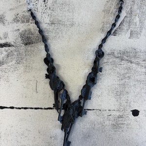 May include: A black necklace featuring a collection of small keys. The necklace has a silver chain and a dark, textured pendant. The keys are intricately detailed, creating a unique and gothic aesthetic. The necklace is displayed against a textured, off-white background.