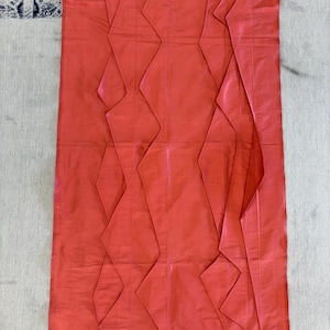Puede incluir: Un panel de tela de color coral vibrante con pliegues en forma de diamante. La tela tiene un brillo sedoso y está orientada verticalmente. El diseño presenta una serie de patrones plegados y angulares, creando una superficie texturizada. El panel es probablemente parte de un proyecto textil más grande.