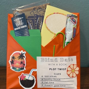 May include: An orange gift box labeled "Blind Date With a Book" with stickers of a sunset, a coconut drink, and a flower. The box contains a Bible emergency numbers card, a yellow card, and a green envelope.