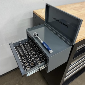 May include: A grey metal U.S. General tool chest with an open lid and an extended drawer containing a set of silver metal sockets. A Quinn brand ratchet with a blue and silver handle rests inside the open top compartment of the tool chest.