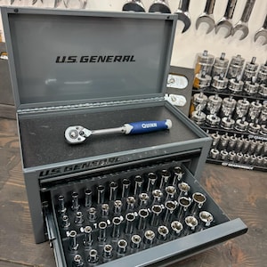 May include: A gray metal tool chest with the text "U.S. GENERAL" on the lid and front. The open drawer reveals a set of sockets. A ratchet wrench with a blue handle, labeled "QUINN", rests inside the chest.