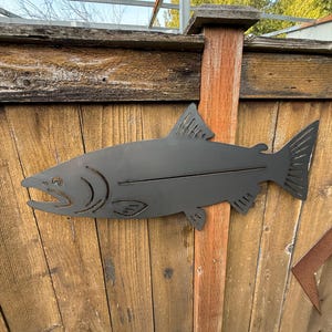 May include: A dark gray metal fish silhouette mounted on a wooden fence. The fish has detailed fins and gills, and is a decorative piece for outdoor spaces. The fence is made of weathered wooden planks.
