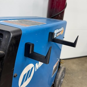 May include: Two black L-shaped hooks mounted on the side of a blue Millermatic 12 Auto-Set welding machine. The hooks are positioned horizontally, one above the other, and feature a pointed upward tip at the end of each arm for cable storage.