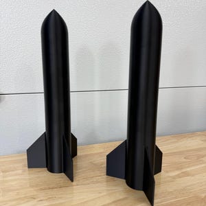 May include: Two matte black rocket-shaped sculptures standing upright. Each object features a smooth, cylindrical body with a pointed, conical top and three flat, triangular fins extending from the base, creating a stable, vertical structure.