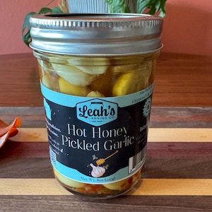 Peut inclure: Un bocal en verre rempli d'ail mariné dans une saumure au miel. Le bocal porte une étiquette bleue et blanche sur laquelle est inscrit "Leah's Canning Co. Hot Honey Pickled Garlic" et "Net Wt. 8oz (227g)".