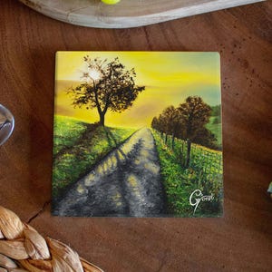 Könnte beinhalten: Quadratische Keramikfliese mit einer Landschaftsmalerei einer Straße, die durch ein Feld auf einen Baum unter einem gelben Himmel führt. Die Fliese ist von einem Holztisch, Trauben, einer Erdbeere, einem Löffel und einem gemusterten Stoff umgeben.