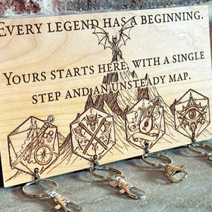 May include: Wooden key holder with engraved text: "Every legend has a beginning. Yours starts here, with a single step and an unsteady map." Features a dragon, mountain, and various symbols. Includes four key rings with clasps.