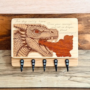 May include: Wooden key holder featuring a detailed dragon illustration with the text "The day calls, hero. Draw your courage and let no dragon stand unchallenged." Includes five black hooks for keys.