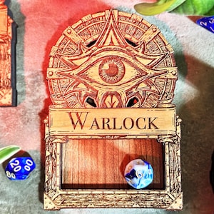 May include: Wooden dice holder with the word "Warlock" engraved on it. The holder features an eye design and a compartment holding a blue and white die. Other dice are scattered around the holder.