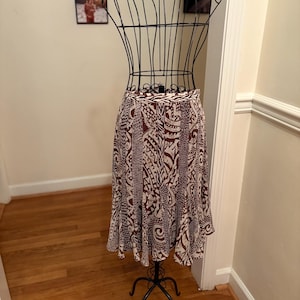 Puede incluir: Una falda hasta la rodilla con un estampado tribal marrón y blanco se exhibe en un maniquí de metal negro. La falda presenta un diseño repetido de formas y líneas abstractas. El dobladillo de la falda tiene un ligero volante.