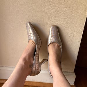 May include: Silver high heels with a square toe and a textured, crocodile-like pattern. The heels have a thin, curved heel. The shoes are positioned on a neutral background.