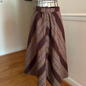 May include: A vintage midi skirt with a brown and striped pattern. The skirt features a high waist and a flared silhouette. The stripes include shades of brown, tan, and a hint of blue. The skirt is displayed on a mannequin.