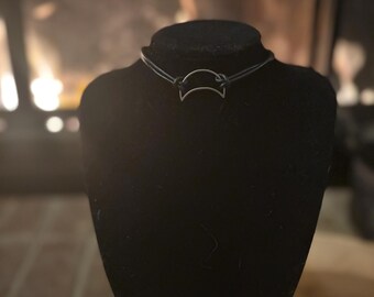 Puede incluir: Collar gargantilla plateado con un colgante de media luna, exhibido en un busto negro. El collar está ensartado en un cordón fino y oscuro. El fondo está borroso, con un brillo cálido y acogedor.