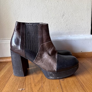 May include: A dark brown and black leather ankle boot with a high block heel and a thick platform sole. The boot features a side elastic gore panel, a rounded toe, and a multi-panel construction with contrasting leather textures and tones.