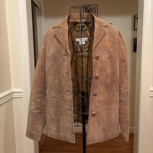 Può includere: Blazer marrone chiaro con motivo a pelle di serpente, con tre bottoni e colletto a revers. La giacca è esposta su un manichino di metallo nero. La fodera interna è di colore marrone chiaro. Il blazer ha due tasche anteriori.