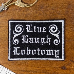 May include: Black and white embroidered patch with the text "Live Laugh Lobotomy". Gold scissors and a white measuring tape are also on a wooden surface.
