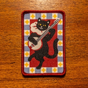 May include: A rectangular embroidered patch featuring a black cat with yellow eyes wearing a white ruffled collar and playing a banjo. The cat is centered on a red background surrounded by a border of light blue and white squares with yellow stars.