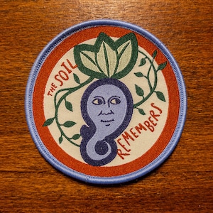 May include: A circular embroidered patch with a light blue border, featuring a central purple figure with a human-like face, green leaves above, and green vines surrounding it. The text THE SOIL REMEMBERS is stitched in red on a cream background.