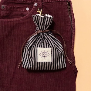 May include: A small drawstring pouch featuring a black and grey vertical striped pattern. The pouch has a brown leather cord closure and a white square patch on the front with a dark embroidered eye design. A gold-toned metal lobster clasp is attached.