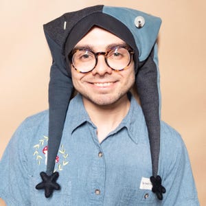 May include: A jester-style hat featuring a two-toned design of dark charcoal gray and muted blue fabric. The hat has two long, pointed side flaps that hang down, each tipped with a black star-shaped ornament. A silver button is attached to the blue side.
