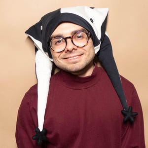 May include: A jester hat featuring a black and white color-blocked design with two long, pointed tails. Each tail ends in a small, black, star-shaped pom-pom. The hat is constructed from fabric and includes small buttons along the side seams.