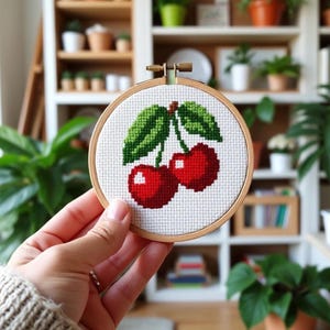 Puede incluir: Un bastidor de bordado de punto de cruz con un par de cerezas rojas con hojas verdes. El diseño está sobre un fondo de tela blanca, enmarcado por un aro de madera. El proyecto de artesanía se sostiene frente a un fondo borroso de plantas y estanterías.