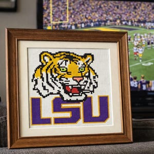 May include: Framed cross-stitch art featuring the LSU Tigers logo. The design includes a yellow and black tiger head above the purple letters "LSU". The artwork is set in a brown wooden frame with a white mat, perfect for a fan's home decor.