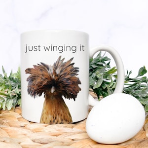 Peut inclure: Une tasse en céramique blanche avec un poulet brun et blanc à la tête volumineuse et duveteuse et le texte "just winging it" imprimé sur le devant.