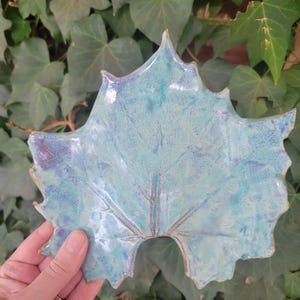 May include: A ceramic leaf-shaped dish with a turquoise glaze and subtle purple accents. The dish has a detailed vein pattern and a glossy finish. The leaf is held against a backdrop of green ivy.