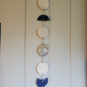 May include: A ceramic moon phase wall hanging. The phases include white circles, a dark blue half-circle with glitter, a crescent moon, and a gray crescent shape. The pieces are connected by silver chains.