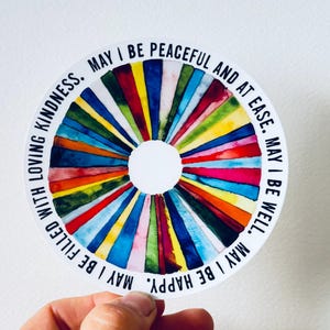 May include: A round sticker with a colorful watercolor design radiating from a white center. The design features vertical stripes in various colors, including red, blue, yellow, and green. The sticker has the text "MAY I BE PEACEFUL AND AT EASE. MAY I BE WELL. MAY I BE HAPPY. MAY I BE FILLED WITH LOVING KINDNESS."