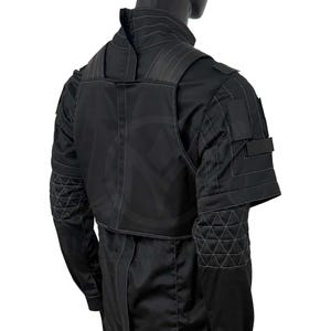 Puede incluir: Chaqueta t&aacute;ctica negra con cuello alto y detalles de costuras geom&eacute;tricas. La chaqueta presenta una superposici&oacute;n tipo chaleco y m&uacute;ltiples bolsillos. El dise&ntilde;o incluye un patr&oacute;n sutil y repetitivo en la espalda y las mangas.