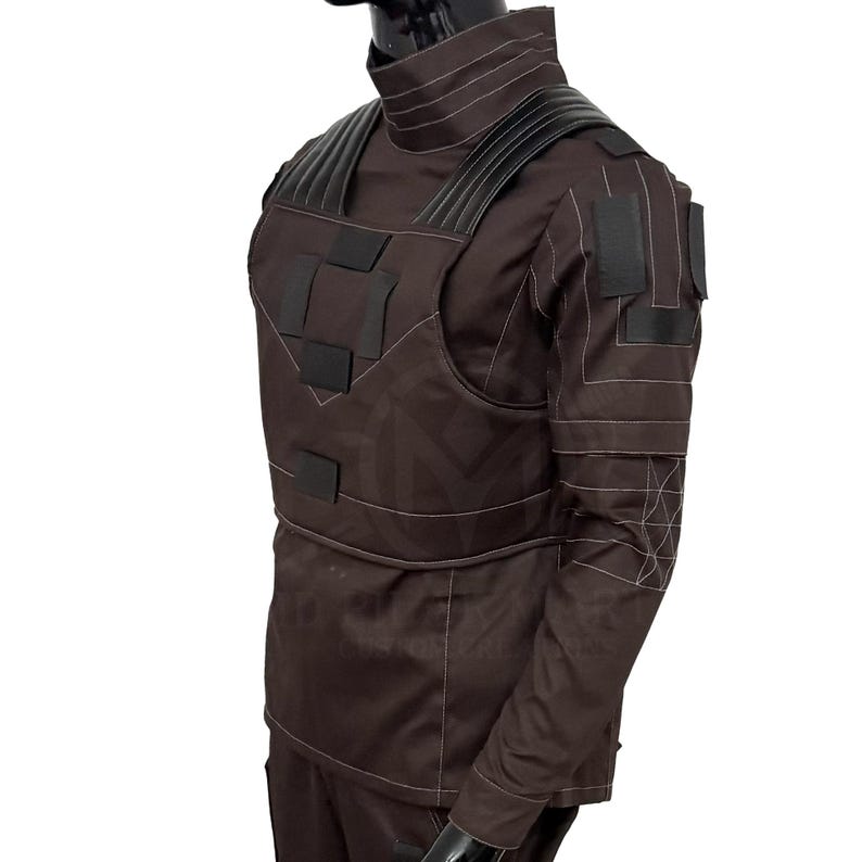 Puede incluir: Un chaleco t&aacute;ctico y uniforme marr&oacute;n con detalles en negro. El chaleco presenta m&uacute;ltiples parches y correas rectangulares, con costuras blancas. El uniforme tiene cuello alto y mangas largas. El conjunto se exhibe en un maniqu&iacute;.
