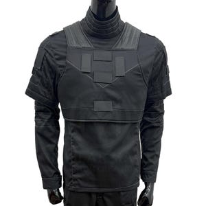 Puede incluir: Chaleco t&aacute;ctico negro con cuello alto y mangas cortas. El chaleco presenta m&uacute;ltiples parches rectangulares y &aacute;reas de hombros reforzadas. La prenda est&aacute; dise&ntilde;ada para la protecci&oacute;n y tiene una apariencia elegante y moderna.