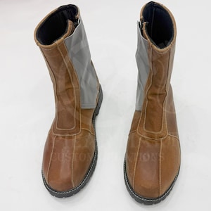 Puede incluir: Un par de botas de cuero marrón con un panel gris y suelas negras. Las botas tienen cierre de cremallera y están diseñadas para llevarse a media pantorrilla. Las costuras son visibles y las botas parecen nuevas.