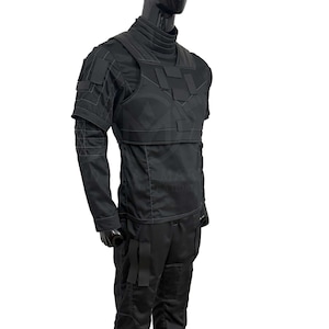 Puede incluir: Chaleco t&aacute;ctico negro y camisa de manga larga. El chaleco presenta m&uacute;ltiples paneles rectangulares y se usa sobre una camisa de cuello alto. Las mangas tienen un dise&ntilde;o geom&eacute;trico. Los pantalones son negros con bolsillos cargo. El maniqu&iacute; es negro.