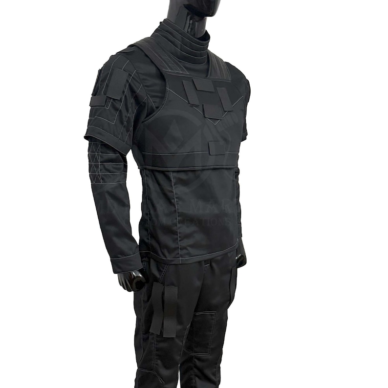 Puede incluir: Chaleco t&aacute;ctico negro y camisa de manga larga. El chaleco presenta m&uacute;ltiples paneles rectangulares y se usa sobre una camisa de cuello alto. Las mangas tienen un dise&ntilde;o geom&eacute;trico. Los pantalones son negros con bolsillos cargo. El maniqu&iacute; es negro.