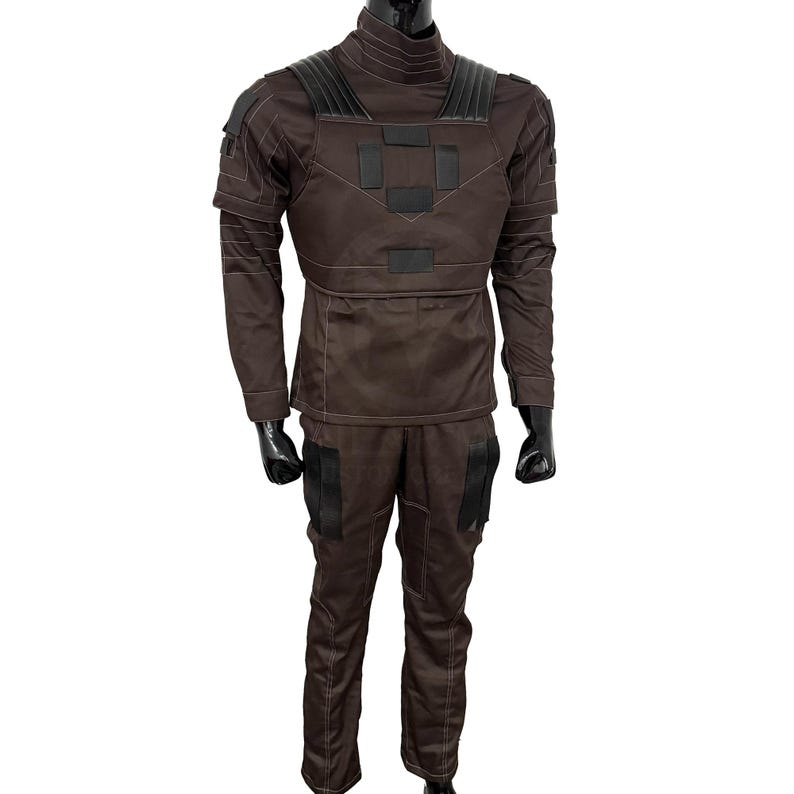Puede incluir: Un conjunto de uniforme t&aacute;ctico marr&oacute;n, que incluye una camisa de manga larga con cuello alto y un pantal&oacute;n a juego. La camisa presenta detalles negros en los hombros y el pecho. Los pantalones tienen bolsillos en los muslos. El conjunto se muestra en un maniqu&iacute;.