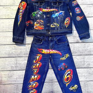 K&ouml;nnte beinhalten: Dunkelblaues Jeansjacken- und Jeans-Set mit Hot Wheels-Motiven. Die Jacke und Jeans sind mit bunten Auto- und Logo-Designs versehen. Der Name "Jamari" ist auf der Jeans aufgedruckt.