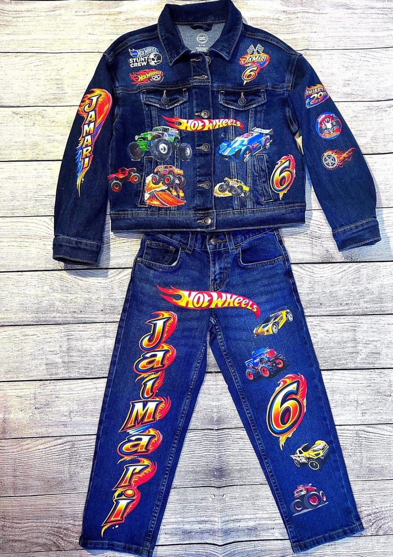 K&ouml;nnte beinhalten: Dunkelblaues Jeansjacken- und Jeans-Set mit Hot Wheels-Motiven. Die Jacke und Jeans sind mit bunten Auto- und Logo-Designs versehen. Der Name "Jamari" ist auf der Jeans aufgedruckt.