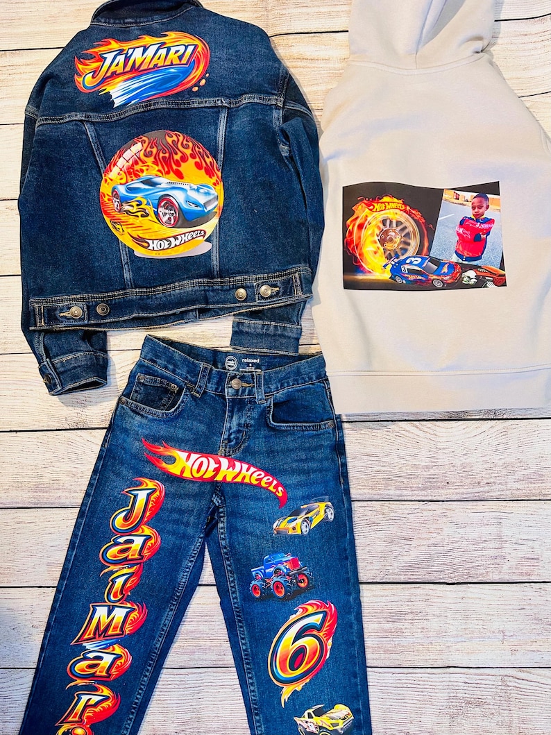 K&ouml;nnte beinhalten: Ein Kinderbekleidungsset mit Jeansjacke, Jeans und Kapuzenpullover mit Hot Wheels-Grafiken. Die Jacke und Jeans tragen den Namen "Jamari" und Hot Wheels-Logos. Der Hoodie hat ein Foto und Hot Wheels-Grafiken. Die Jeans hat die Nummer "6".