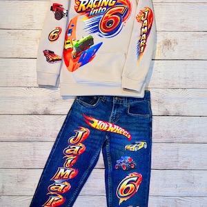 K&ouml;nnte beinhalten: Ein beiges Kapuzenshirt und eine blaue Jeans-Kombination mit Rennsport-Motiv. Das Kapuzenshirt zeigt "Racing into 6" Grafiken mit Rennwagen. Die Jeans hat "Hot Wheels" und "Jamari" Schriftz&uuml;ge sowie Autografiken. Ein lustiges Outfit f&uuml;r ein Kind.