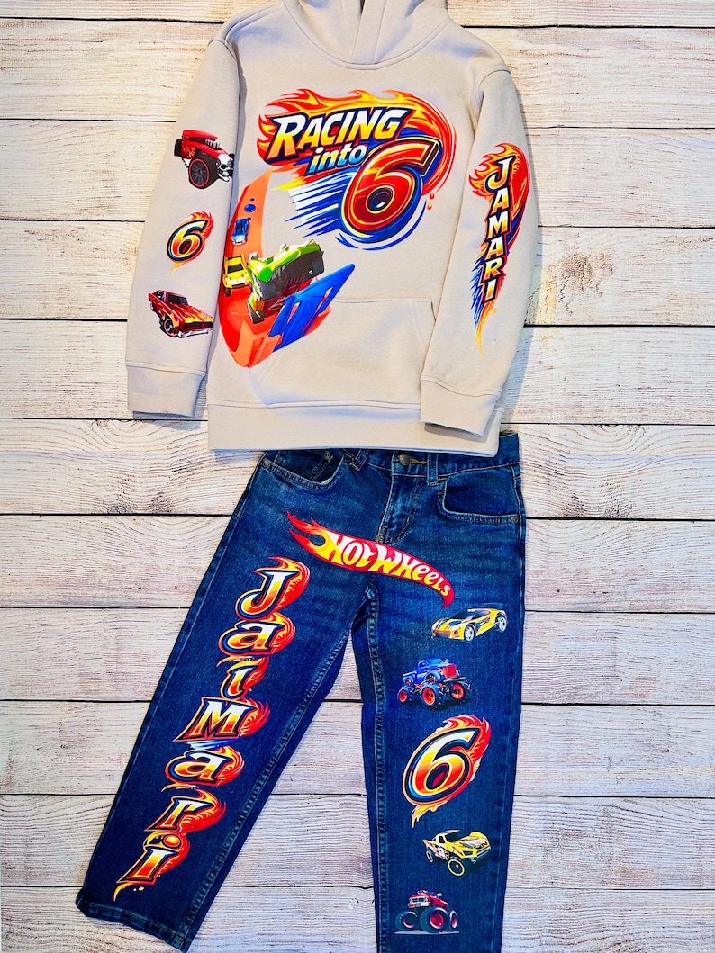 K&ouml;nnte beinhalten: Ein beiges Kapuzenshirt und eine blaue Jeans-Kombination mit Rennsport-Motiv. Das Kapuzenshirt zeigt "Racing into 6" Grafiken mit Rennwagen. Die Jeans hat "Hot Wheels" und "Jamari" Schriftz&uuml;ge sowie Autografiken. Ein lustiges Outfit f&uuml;r ein Kind.