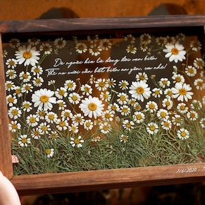 May include: A wooden frame with a glass front containing pressed white daisies and green foliage. The frame has a white inscription that reads "Da ngo&agrave;i kia l&agrave; dang đọc van bila The na cước vẫn biết biến an ytor, com nhe!" and the date "7/6/2020 - 7/6/2024".