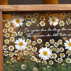 May include: A framed pressed flower art piece with white daisies and green stems. The quote "D&ugrave; ngo&agrave;i kia l&agrave; d&ograve;ng đời vạn biển Th&igrave; nụ cười vẫn bất biến an y&ecirc;n!" is written in white on a brown background.