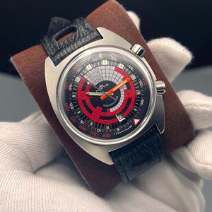 Puede incluir: Un reloj de pulsera de acero inoxidable con una correa de cuero negro. La esfera del reloj es negra con detalles en rojo y blanco, y una aguja naranja. La esfera del reloj tiene el texto "FURTIS MARINEMASTER" y "DEPTH IN FEET".
