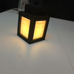 May include: A black, cube-shaped lantern with a warm yellow glow emanating from its rectangular panels. The lantern has a boxy design and sits on a white surface, creating a contrast with the dark fixture.