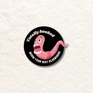 Puede incluir: Una pegatina circular en blanco y negro con un gusano rosa con una expresión de sorpresa. El texto en la pegatina dice "Totally booked, worm your way elsewhere."