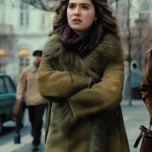 May include: A woman wearing a long olive green coat with a fur collar and a patterned scarf. The coat is belted at the waist. The background shows a city street with pedestrians and buildings.
