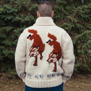 May include: Cream-colored knit sweater featuring a large, detailed design of two foxes in shades of brown and white on the back. The sweater has a ribbed collar and cuffs, with a small floral pattern near the bottom hem. The sweater is worn by a person.